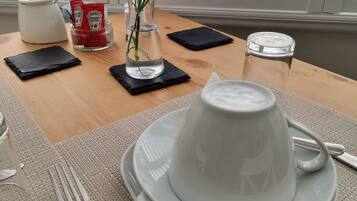 Mesa posta com um bule branco, uma chávena branca, um garfo e um guardanapo. Há um vaso com flores cor-de-rosa e um copo de água sobre a mesa.