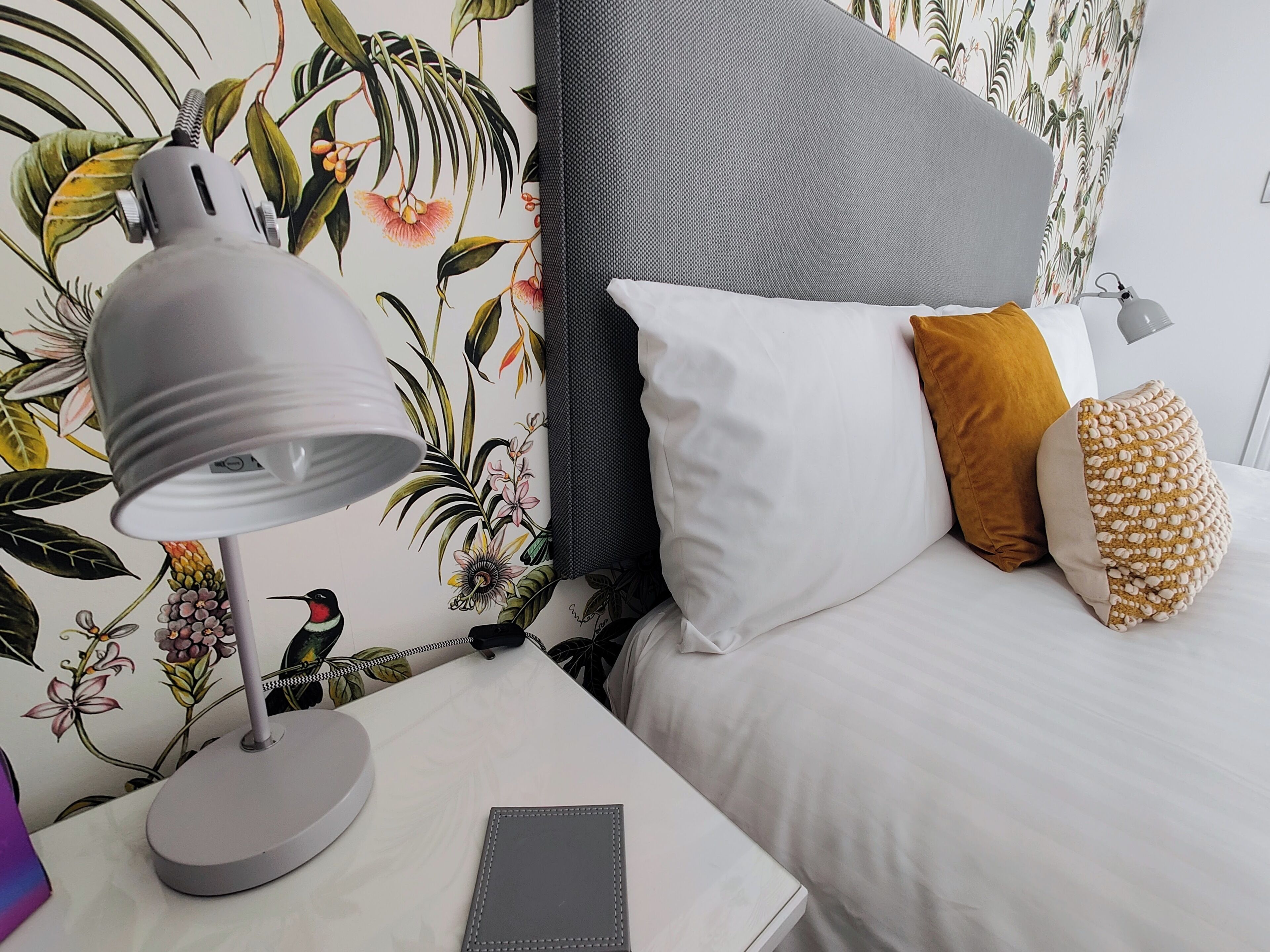 Cama bem arrumada com almofadas brancas e mostarda, abajur cinza em uma mesa de cabeceira e papel de parede floral.