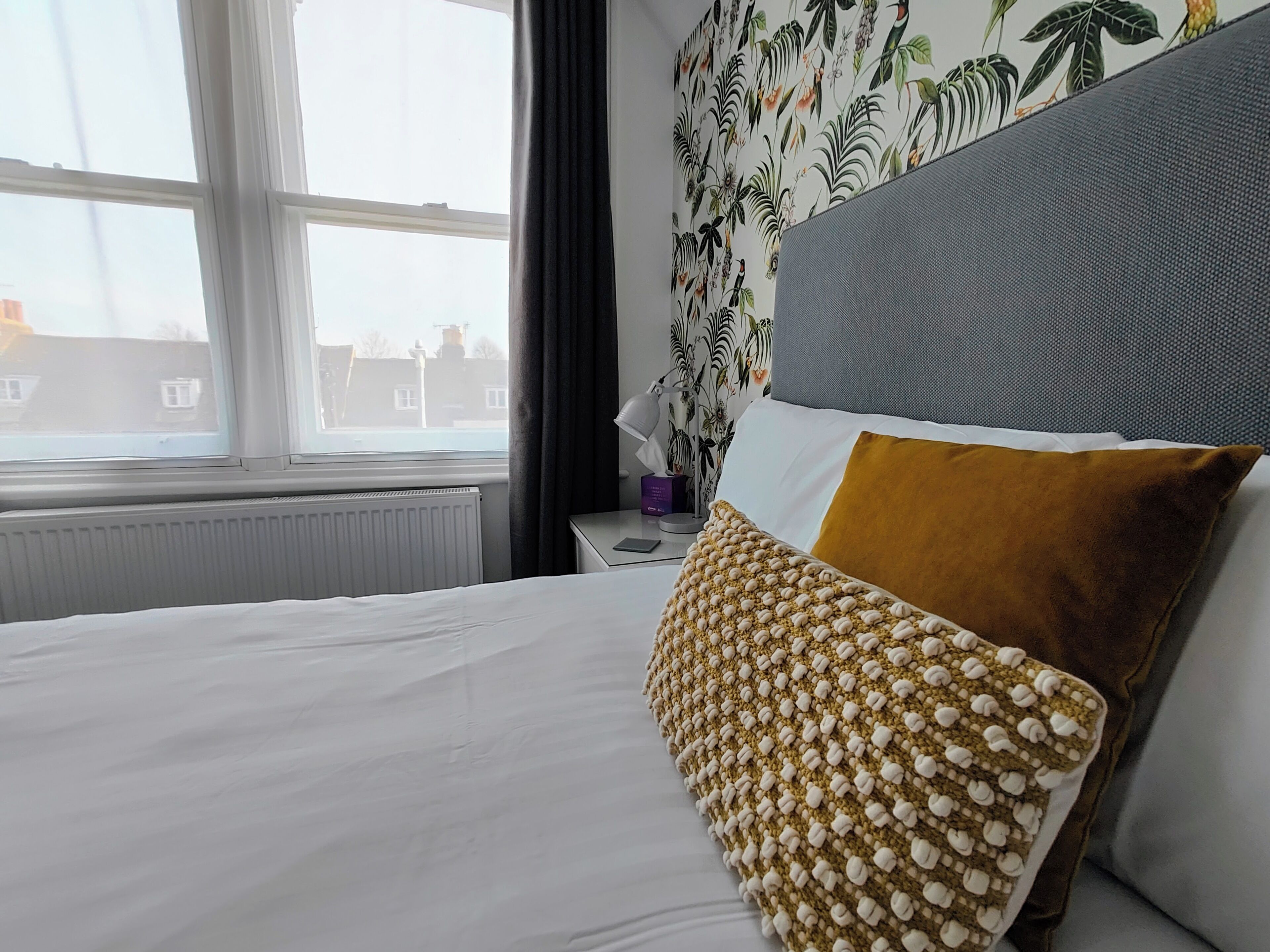 Hotelkamer met een bed, een raam, een radiator, een nachtkastje en een muur met tropische behang.
