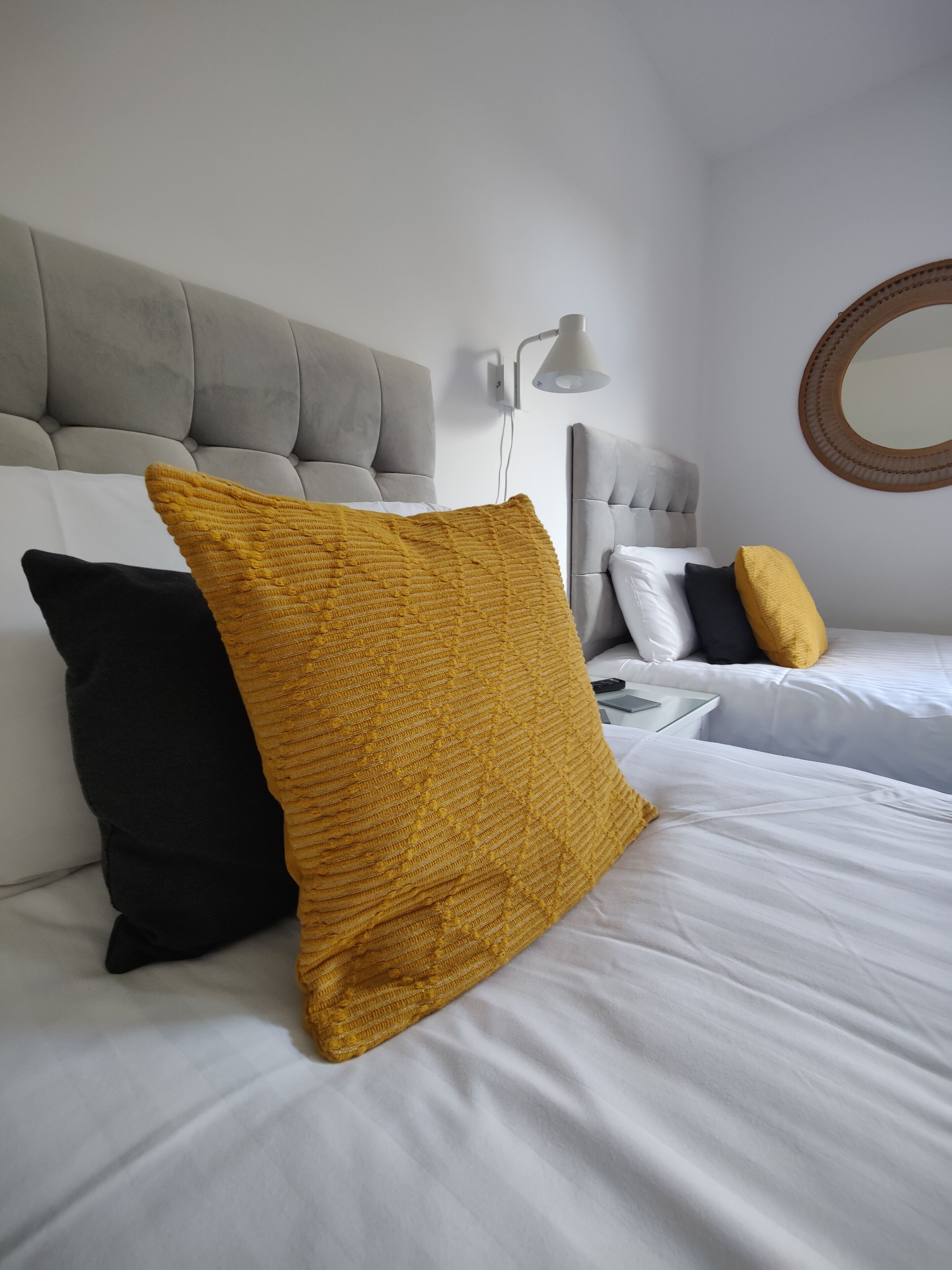 Una cama bien hecha con ropa blanca y dos almohadas, una amarilla y otra negra, en una habitación moderna con un cabecero capitonado y un espejo redondo en la pared.