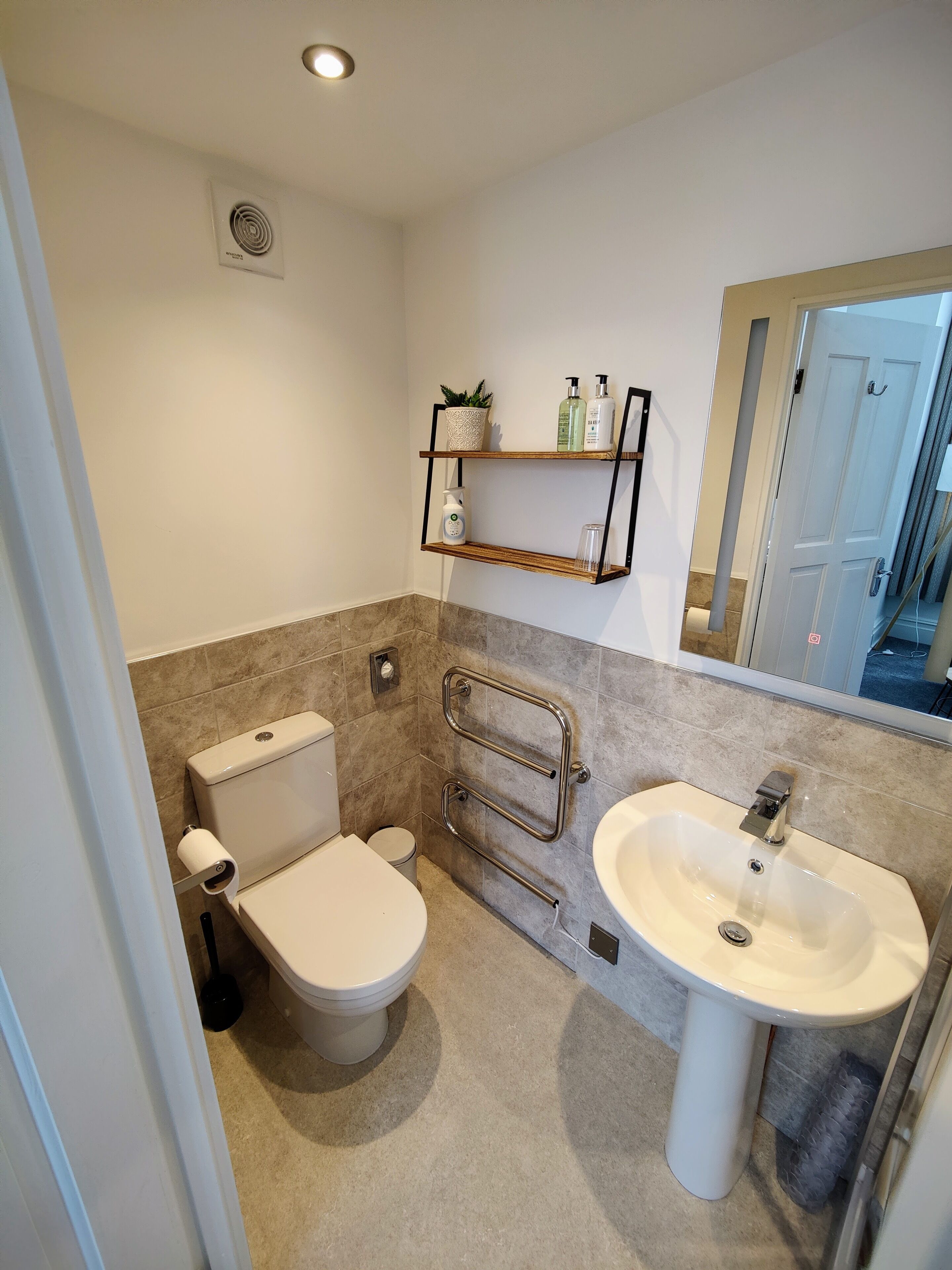 Un baño moderno con inodoro, lavamanos y ducha. Hay repisas con artículos de tocador y una planta en maceta.