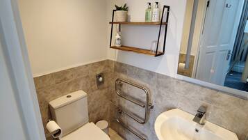 Un baño moderno con inodoro, lavamanos y ducha. Hay repisas con artículos de tocador y una planta en maceta.