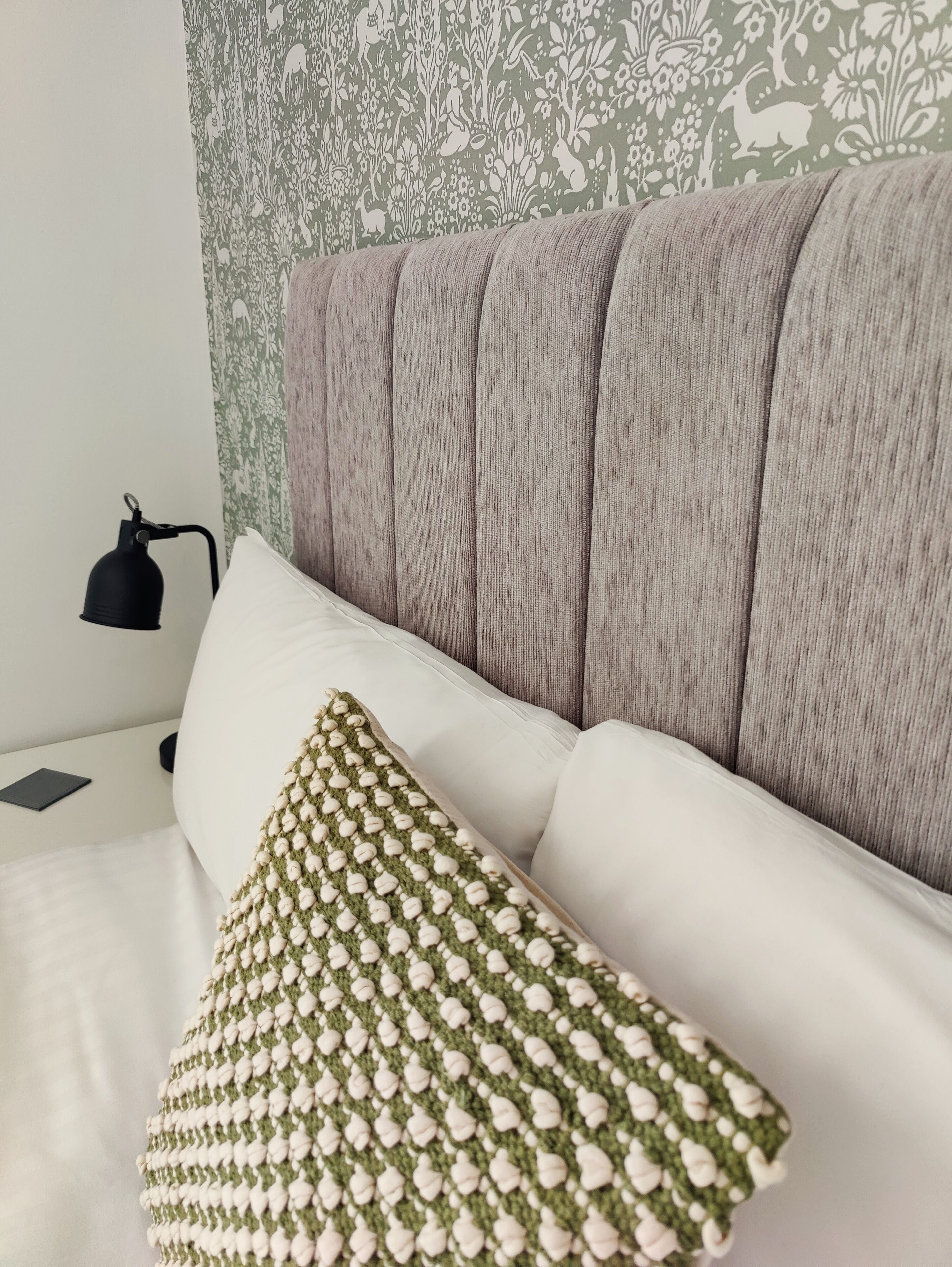 A neatly made bed with a grey upholstered headboard, a white pillow, and a green and white patterned pillow. A black lamp is on the bedside table.
