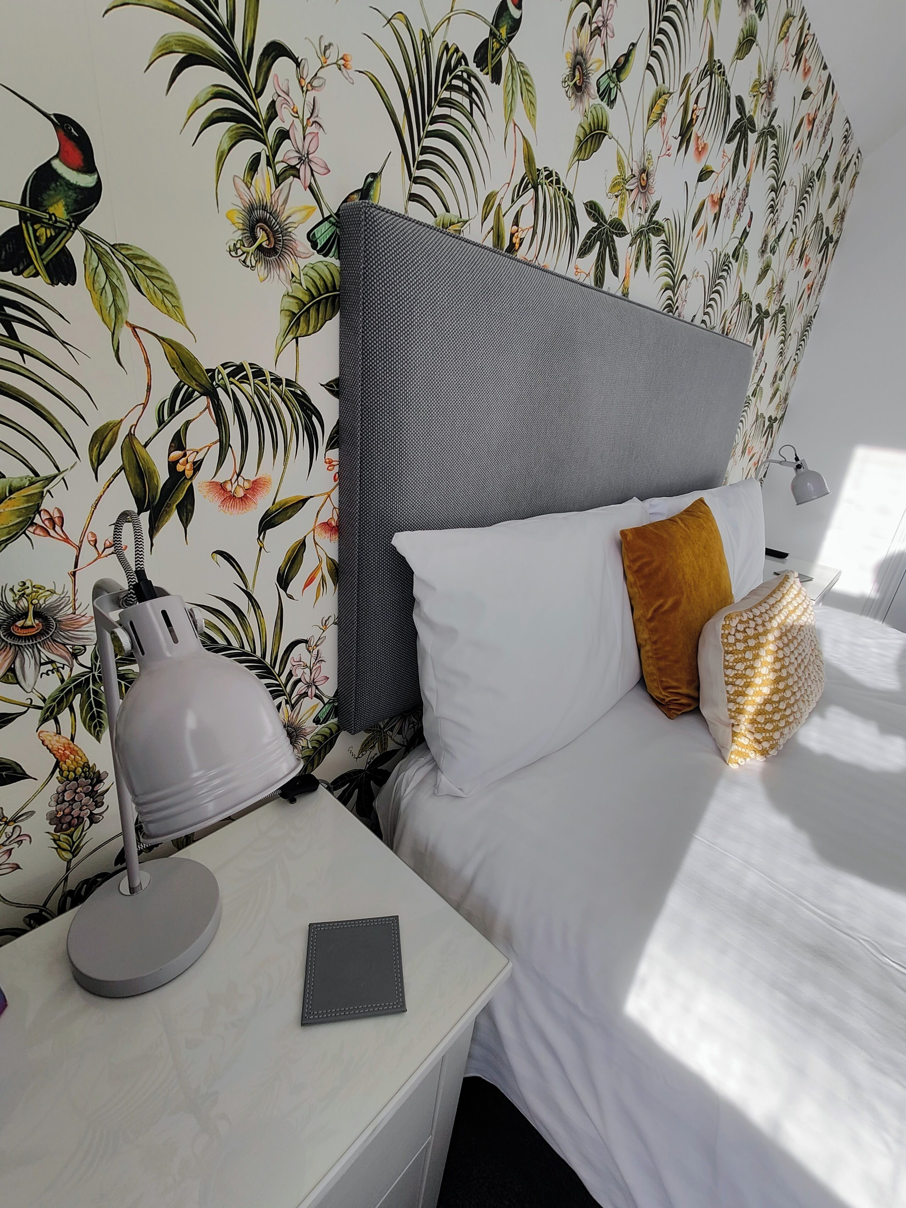 A bedroom with a bed, white bedding, a grey headboard, a bedside lamp, a bedside table with a tablet, and a wall with a tropical wallpaper.