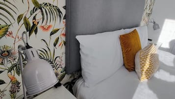 A bedroom with a bed, white bedding, a grey headboard, a bedside lamp, a bedside table with a tablet, and a wall with a tropical wallpaper.