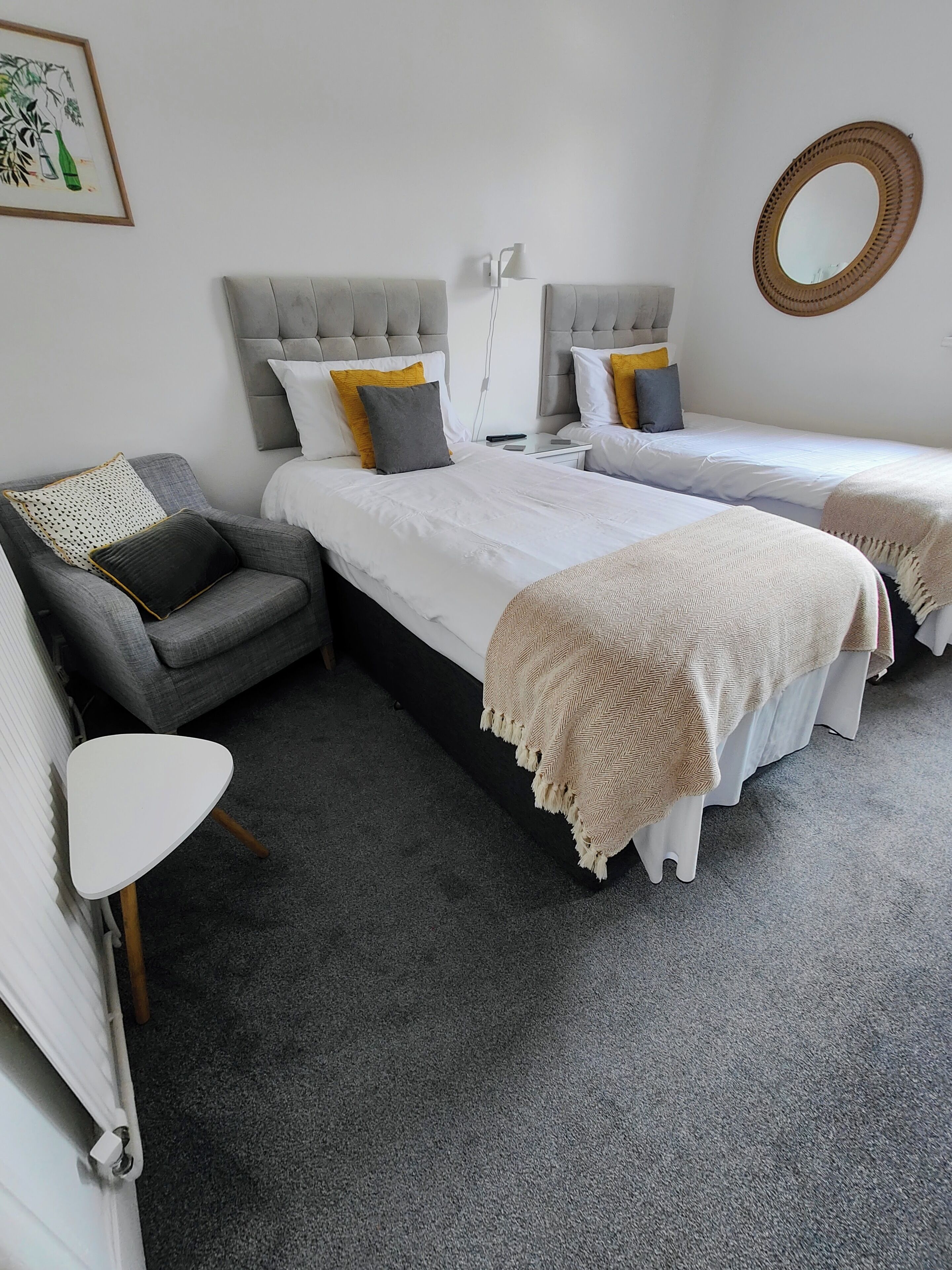 A bedroom with two beds, a grey armchair, a small white table, and a mirror on the wall.