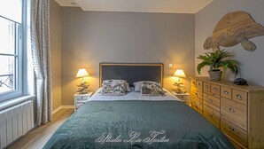 Un dormitorio con una cama, dos mesitas de noche, un aparador, una ventana con cortinas y una planta sujeta a la pared.