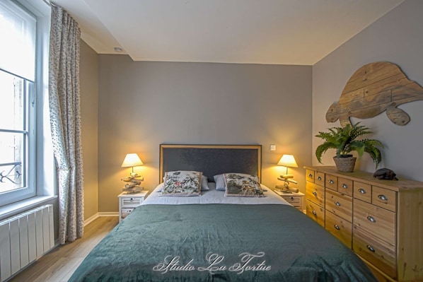 A bedroom with a bed, two bedside tables, a dresser, a window with curtains, and a wall-mounted plant.
