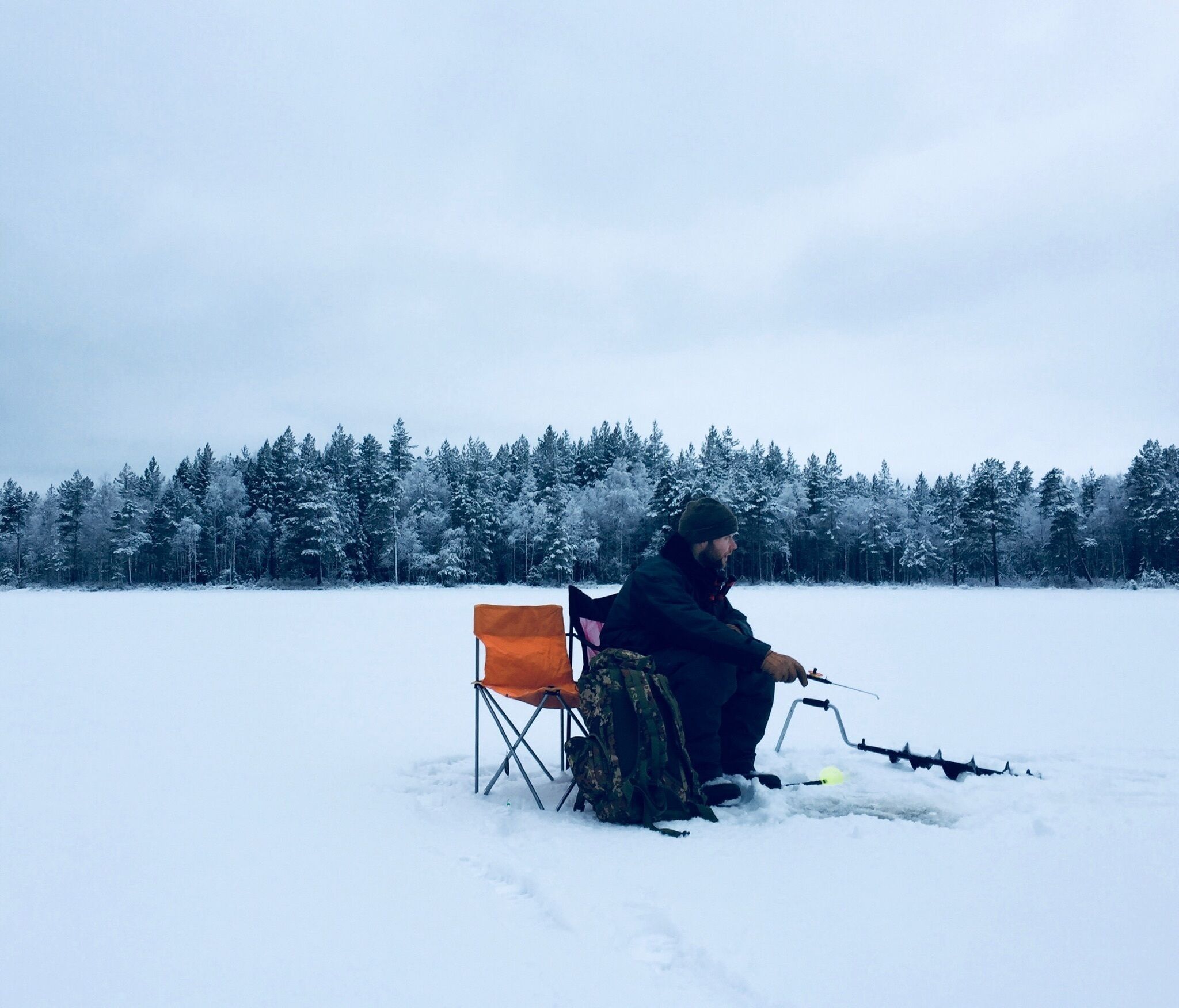 A person sitting on a folding chair in a snowy outdoor setting with a fishing rod.