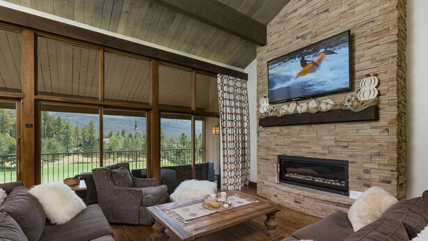 A spacious living room with a large TV mounted on a stone wall, a fireplace, and a view of a golf course through large windows. - Majestic Mountain Retreat | AC, Pool, Hot Tub, Golf & Ski (Durango)