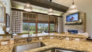 A kitchen with granite countertops, a stainless steel sink, and a dishwasher. There are three pendant lights, a large window with a view of trees, and a fireplace with a TV above it. - Majestic Mountain Retreat | AC, Pool, Hot Tub, Golf & Ski (Durango)