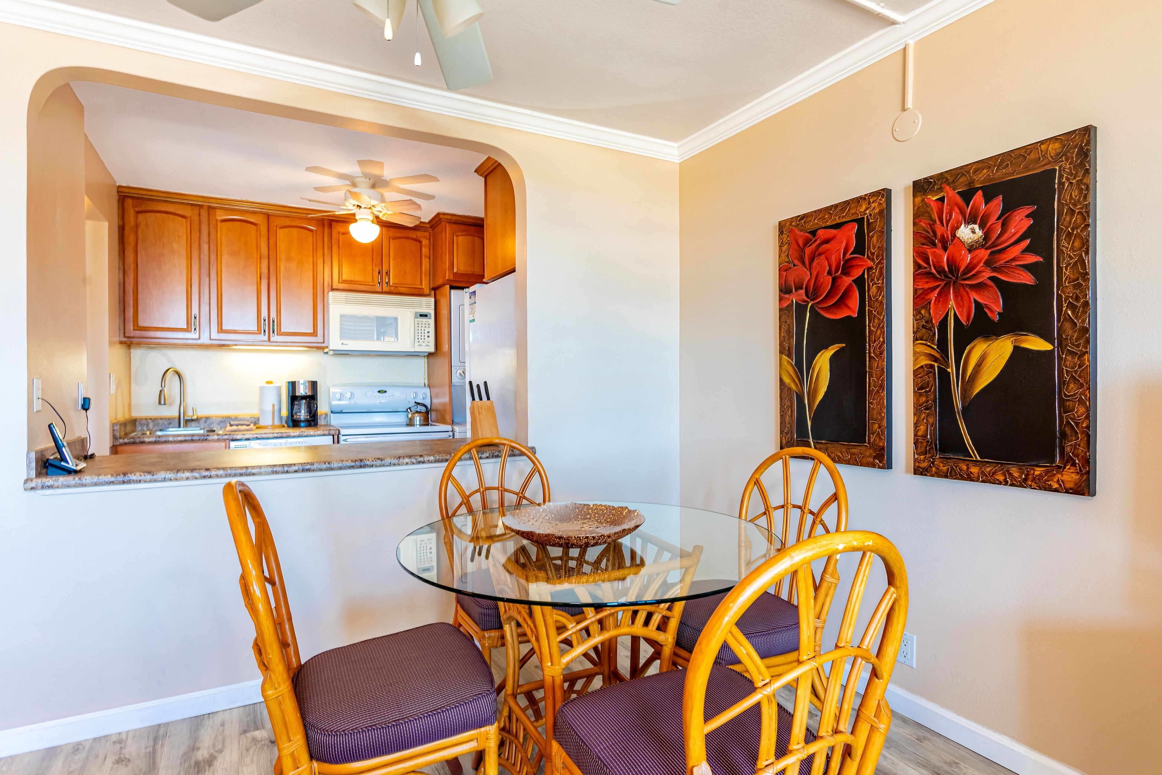 Un comedor con una mesa de vidrio rodeada de sillas de mimbre, una cocina al fondo y dos cuadros con motivos florales en la pared.