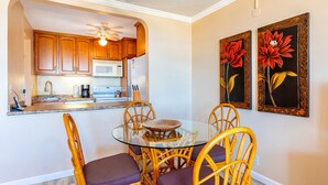 Uma sala de jantar com mesa de vidro rodeada por cadeiras de vime, cozinha ao fundo e duas pinturas florais na parede.