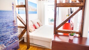 A bedroom with a wooden truss ceiling, a bed with multiple pillows, a small table with a potted plant, and a view of the sea through a window.