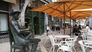 Una statua raffigura un uomo seduto con un libro, tavolini e sedie di un caffè all'aperto, e un edificio verde con decorazioni elaborate sullo sfondo.