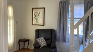 A living room with a black armchair, a small table, a framed picture on the wall, and a staircase leading to another level.
