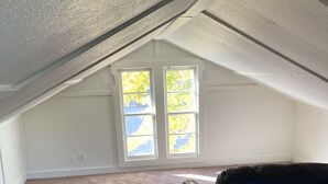 An attic room with a carpeted floor, a black leather couch, and two windows with a view of trees.