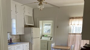A compact kitchen with white cabinetry, a wooden floor, a ceiling fan, and a window with curtains.