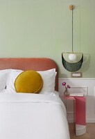 Una cama con ropa de cama blanca y una almohada color mostaza, una mesita de noche rosa con un jarrón de flores, y una lámpara de pared.
