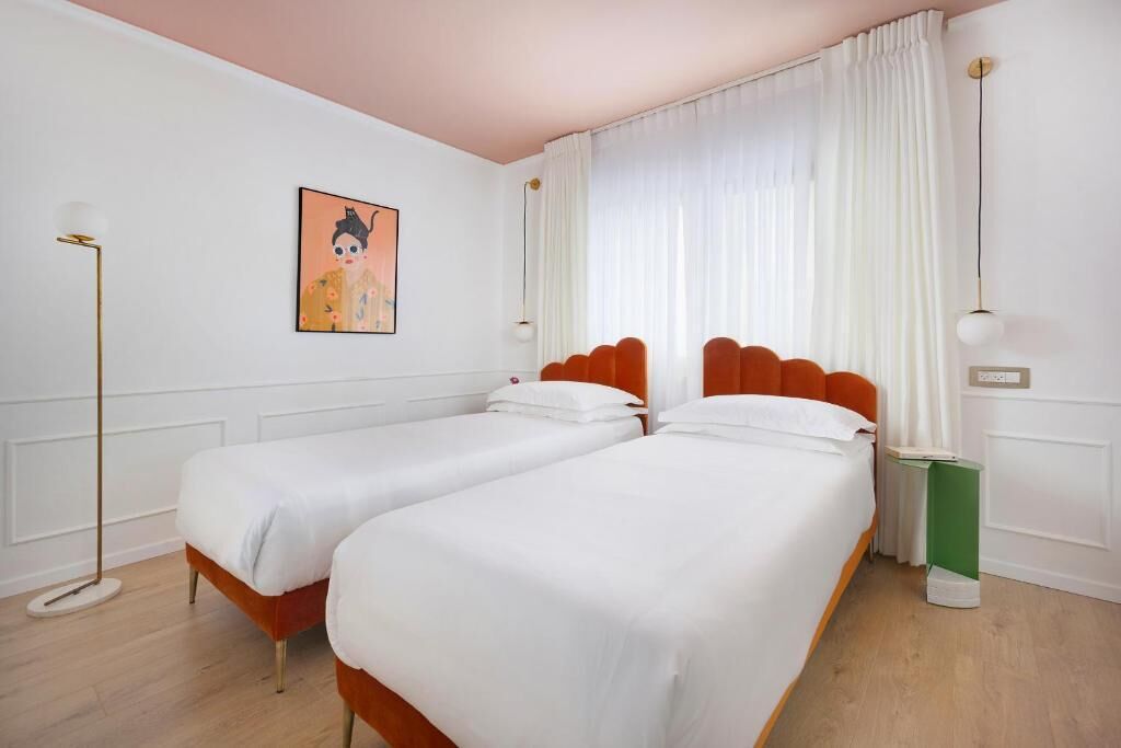A hotel room with two single beds, a large window with white curtains, a floor lamp, and a framed picture on the wall.