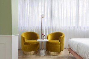 Une pièce avec deux fauteuils de couleur moutarde, une petite table ronde surmontée d’un vase blanc et un lampadaire. On aperçoit un lit avec une literie blanche à l’arrière-plan.