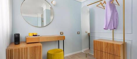 A modern bathroom with a wooden vanity, a round mirror, a towel rack with clothes, and a yellow stool.