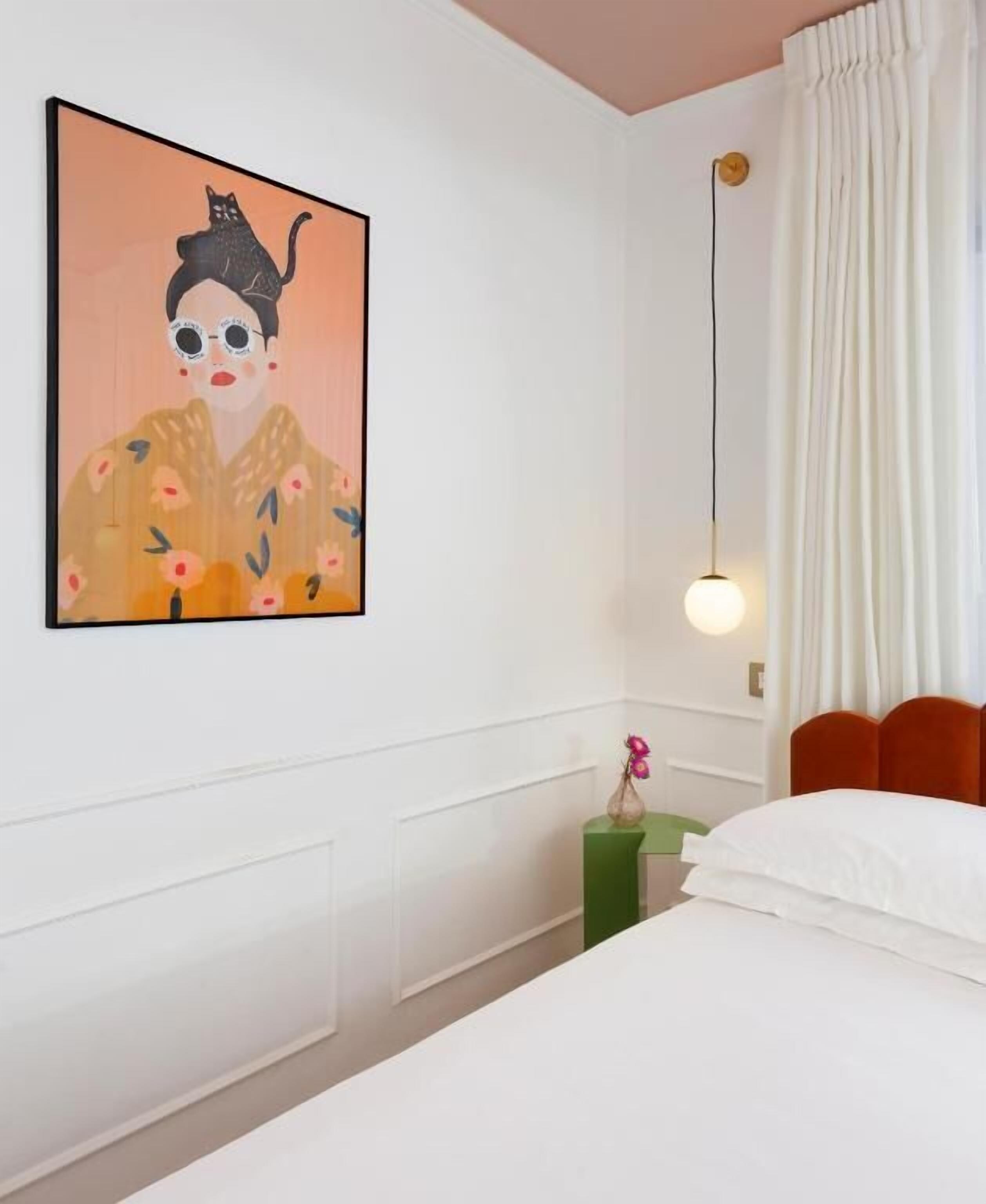 A hotel room with a bed, a nightstand, a vase with a pink flower, a painting on the wall, and a hanging light fixture.