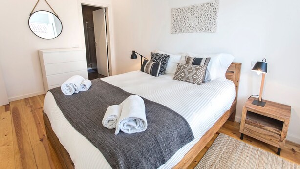 Una cama bien hecha con una manta gris, almohadas blancas y toallas dobladas. Hay una mesita de noche con una lámpara, un revistero de madera y un adorno en la pared sobre la cama.