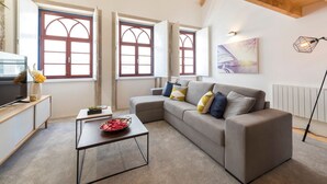 Un soggiorno moderno con un divano grigio, un tavolino da caffè e una TV su un mobile in legno. Ampie finestre con cornici rosse lasciano entrare la luce naturale.