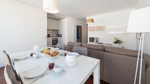 Ett modernt, väl upplyst matområde med ett vitt bord dukat för två personer, en soffa och en tv i bakgrunden.