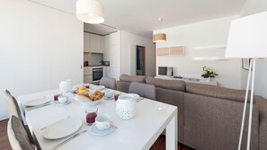 A modern, well-lit dining area with a white table set for two, a sofa, and a TV in the background.
