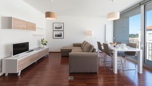 Ett modernt vardagsrum med en platt-TV, en soffa, ett matbord med stolar och stora fönster som erbjuder utsikt över byggnader och grönområden.