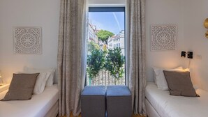 Une chambre d’hôtel avec deux lits, un balcon donnant sur des immeubles et des arbres, et des panneaux décoratifs sur les murs.