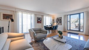 Un salon moderne avec un canapé, un fauteuil et une table basse. On y trouve également un coin repas avec des chaises et une table, ainsi que de grandes fenêtres offrant une vue sur la ville.
