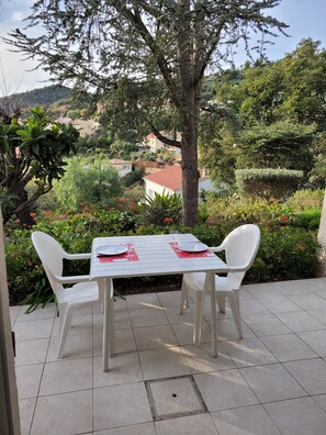 Une terrasse extérieure avec une table et des chaises blanches, entourée de verdure et offrant une vue sur une colline.