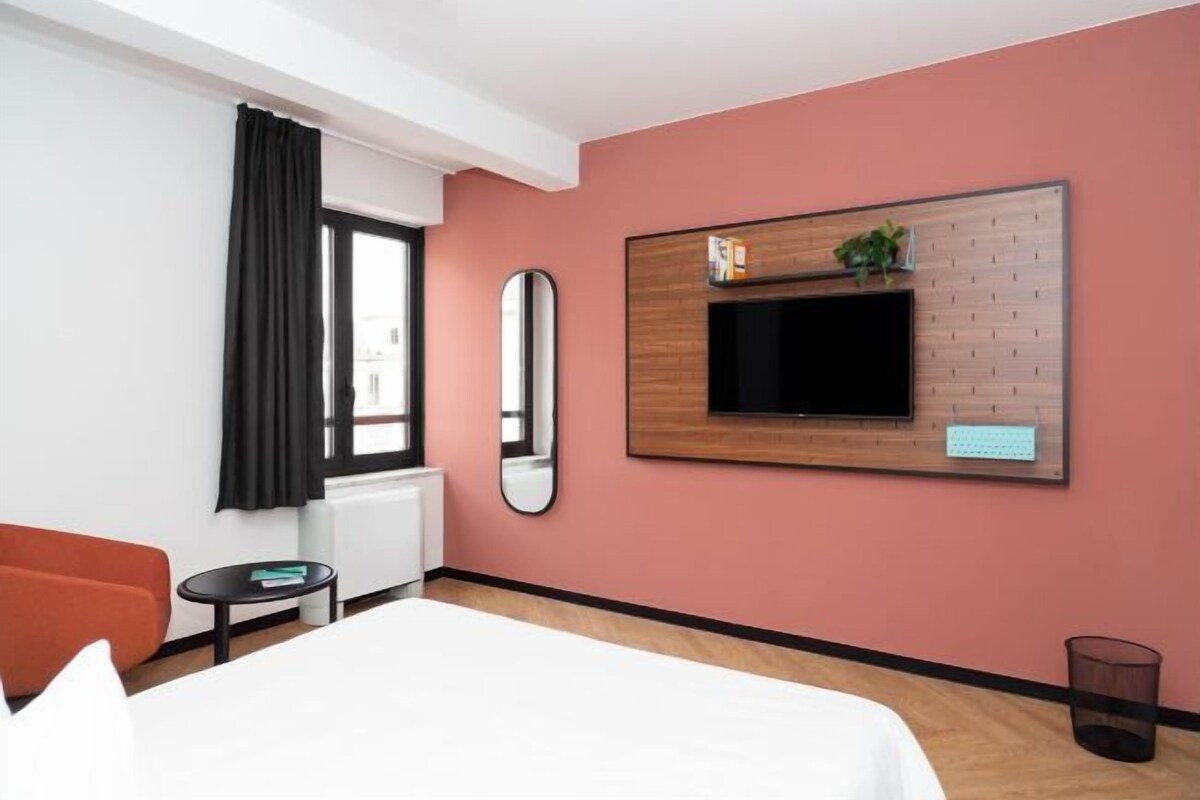 A modern hotel room with a bed, a TV mounted on the wall, a small table, and a window with curtains.