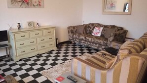 Ein Wohnzimmer mit Schachbrettmuster-Boden, einer Couch, einem gestreiften Sessel, einem kleinen Tisch mit Fernbedienung und einer weißen Kommode mit Schubladen.
