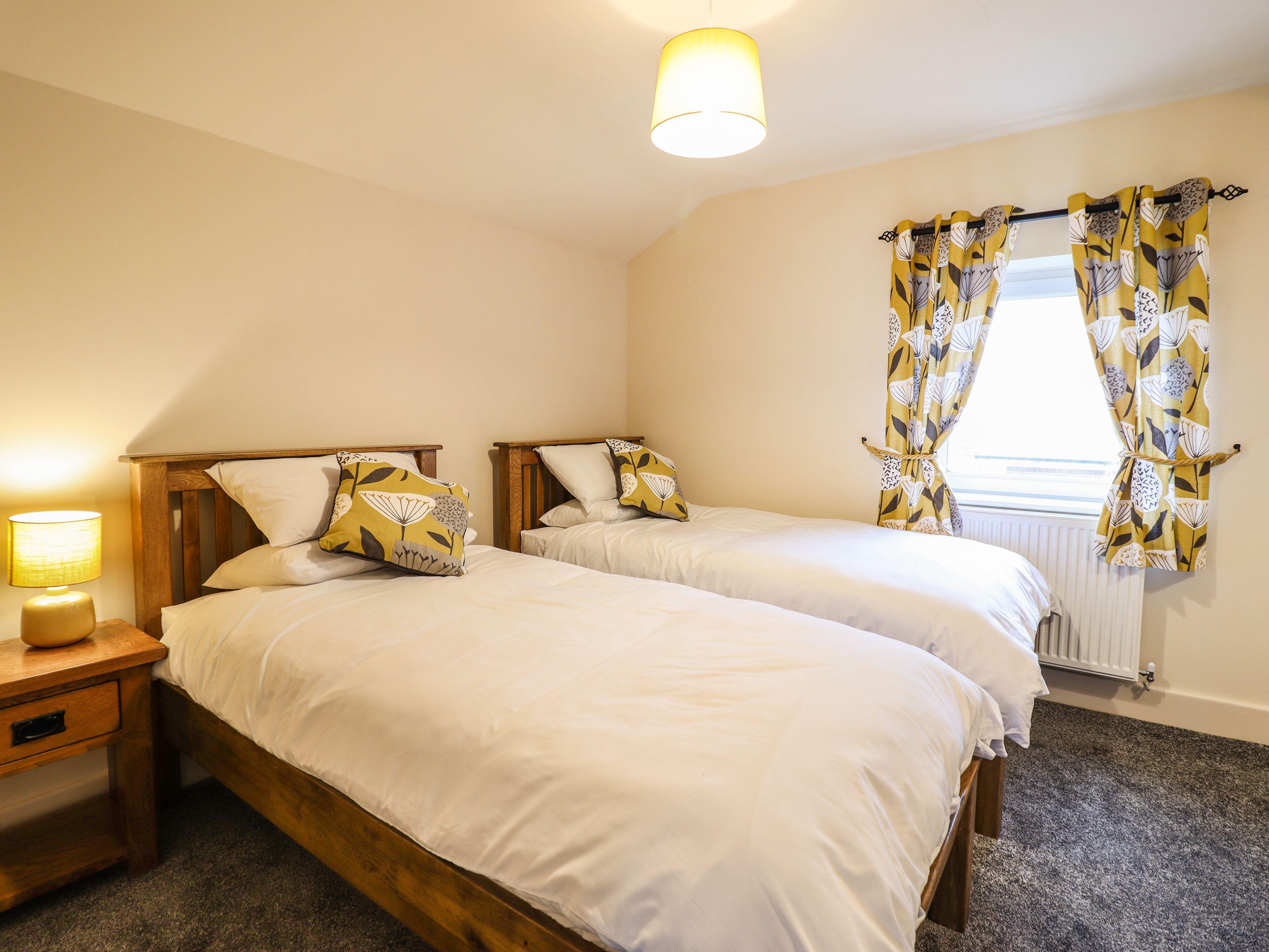 A bedroom with two single beds, a bedside table with a lamp, and a window with curtains.