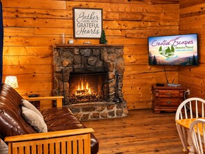 A cozy cabin interior with a stone fireplace, a television displaying a scenic view, and a sign above the fireplace. - Charming 1-Bedroom Log Cabin – Perfect for Family Getaways! (Broken Bow)