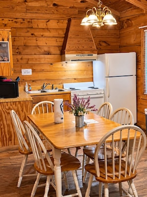 A wooden cabin interior with a dining table, chairs, a kitchen area including a stove and refrigerator, and a hanging light fixture. - Charming 1-Bedroom Log Cabin – Perfect for Family Getaways! (Broken Bow)