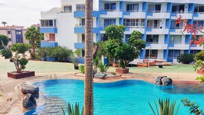 A swimming pool surrounded by white pebbles, palm trees, and a multi-story building with balconies. - Tropical Green Park - Pool - Parking - La Corbeta Building (San Miguel de Abona)