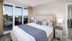 A hotel room with a large bed, a nightstand with a lamp, and a view of the beach through the balcony doors. - Nautilus 2311  Gulf View 2BD/2BA 3rd Floor.  Free Beach Service 3/1-10/31 (Fort Walton Beach)