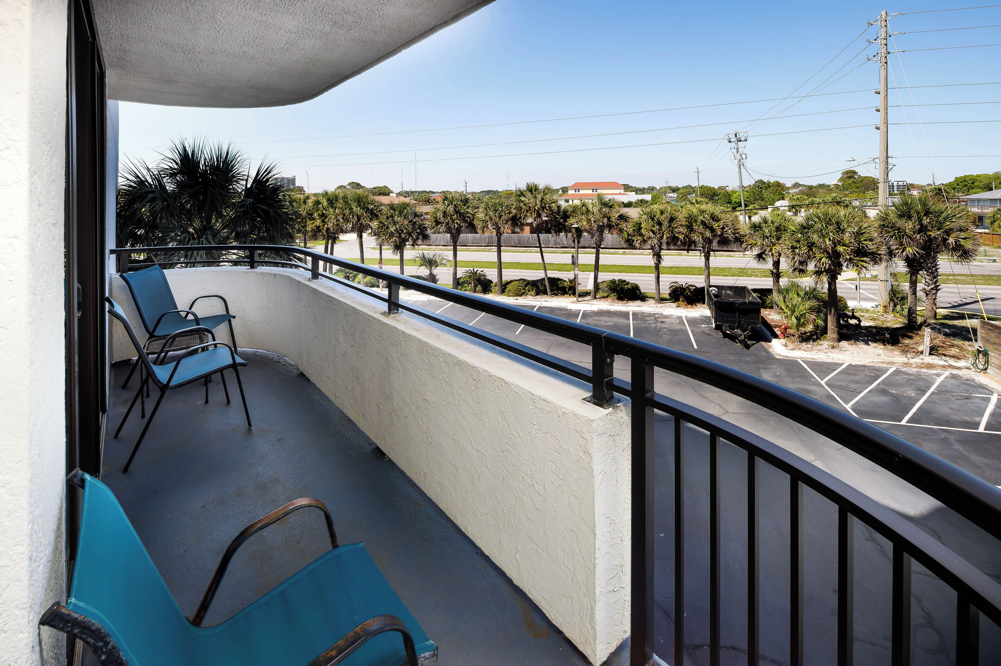 Een balkon met twee blauwe stoelen, een betonnen muur en uitzicht op een parkeerplaats met palmbomen en een gebouw op de achtergrond.