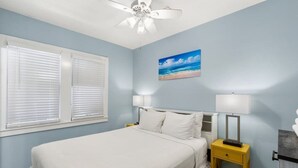Un dormitorio con una cama, dos mesitas de noche, un ventilador de techo, una ventana con persianas y un cuadro con motivos playeros en la pared.