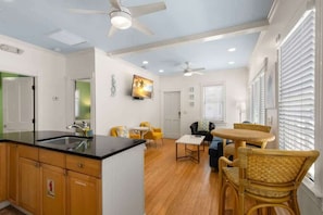 A modern kitchen with a black countertop and wooden cabinets, a dining area with wicker chairs and a small table, and a ceiling fan. - Poolside Paradise - Heated Pool & Steps to Duval St (Key West)
