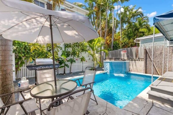 A backyard with a swimming pool, a grill, and a dining area with chairs and an umbrella.