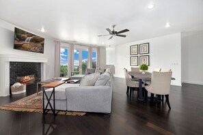 Ein modernes Wohnzimmer mit Kamin, einer Couch, einem Esstisch mit Stühlen und einem Deckenventilator.