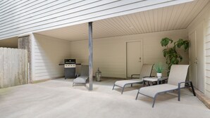 Un patio techado al aire libre con piso de concreto, una parrilla, una silla, una mesita y una planta en maceta.