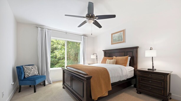 Un dormitorio con cama, mesita de noche, una silla, ventilador de techo y ventana con cortinas.