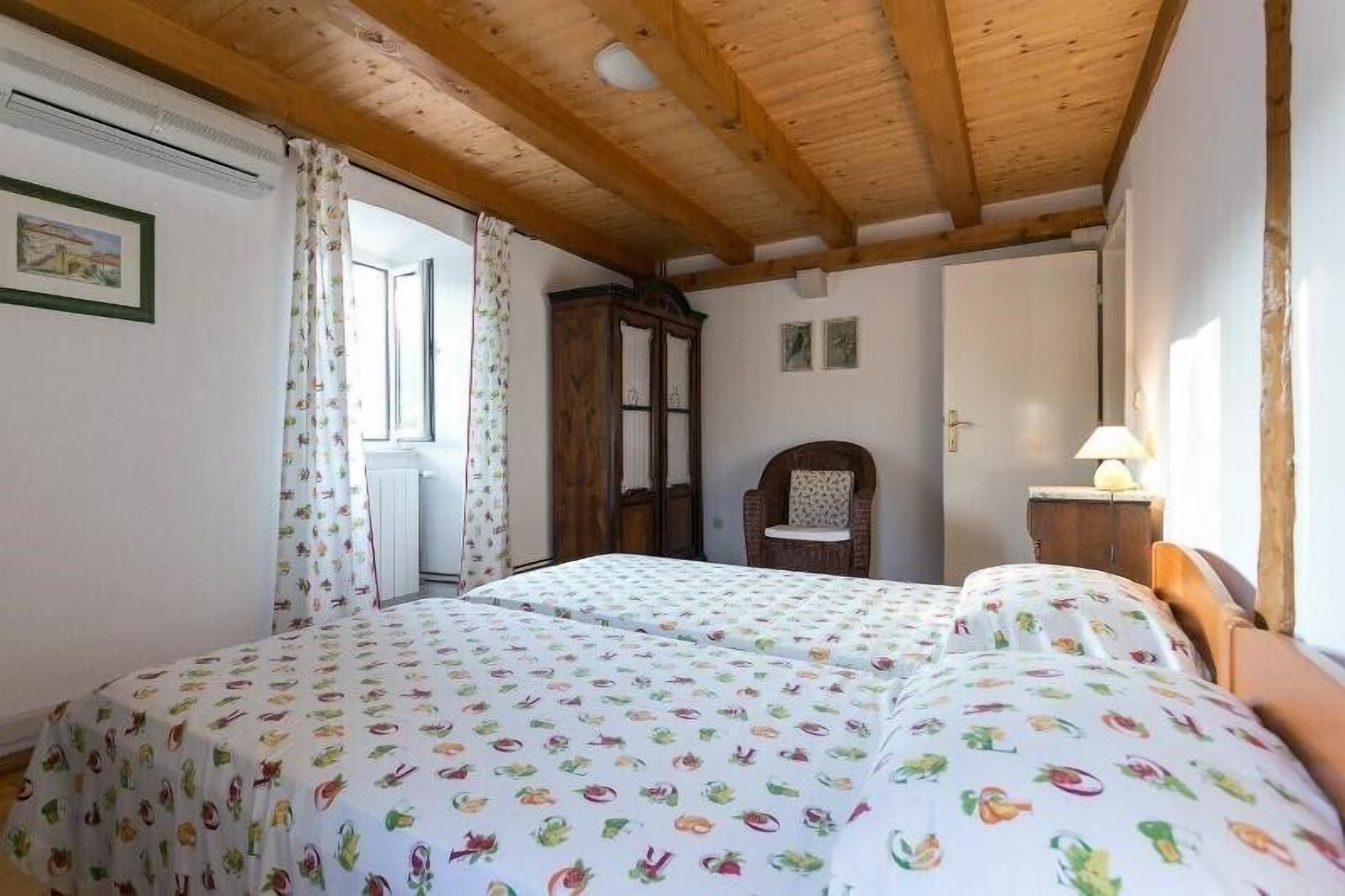 A bedroom with two beds, wooden ceiling beams, a window with curtains, a wooden wardrobe, and a chair.